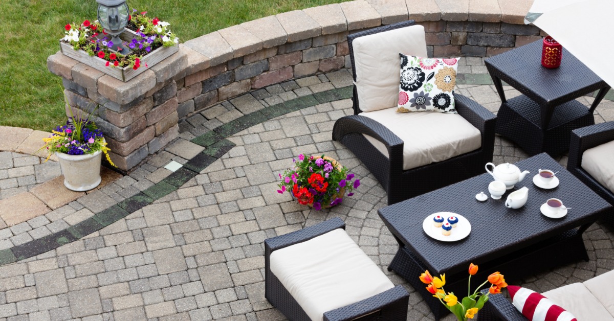 A brick paved patio in a residential yard during the daytime. Two tables and four chairs are setup on the patio.