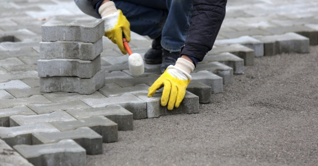 A person wearing white-and-yellow gloves installing patio pavers outside. They are using a mallet to install the pavers.
