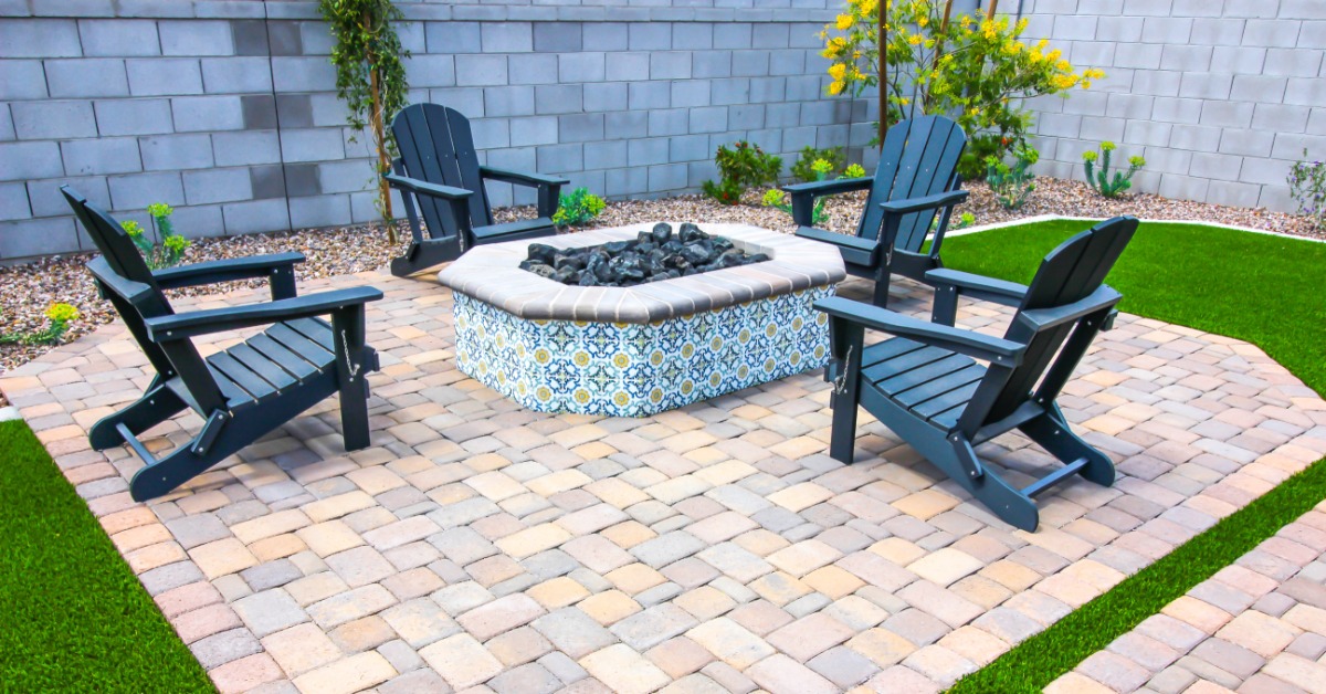 Four black chairs surrounding a fire pit in a residential backyard. The fire pit area is setup on stone pavers.