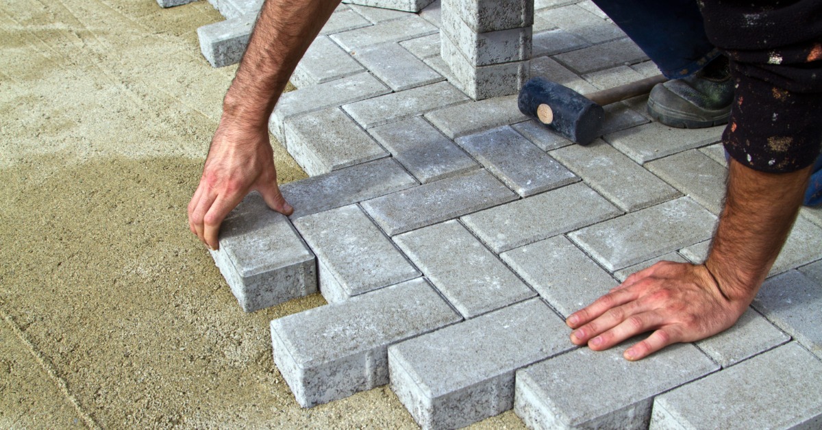 A person using their bare hands to place concrete pavers on a stable bed of sand. A mallet sits next to the installer.