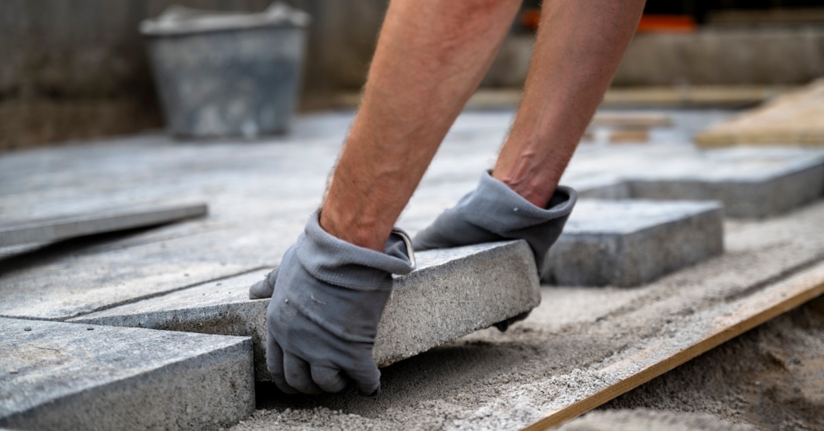 A contractor wearing light blue gloves installing large gray patio pavers outside. A gray bucket is on the pavers.