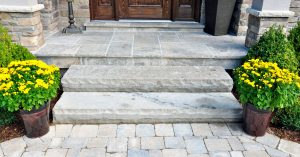 Neutral-toned patio pavers leading up to the gray stone steps in front of a home’s wooden exterior door.