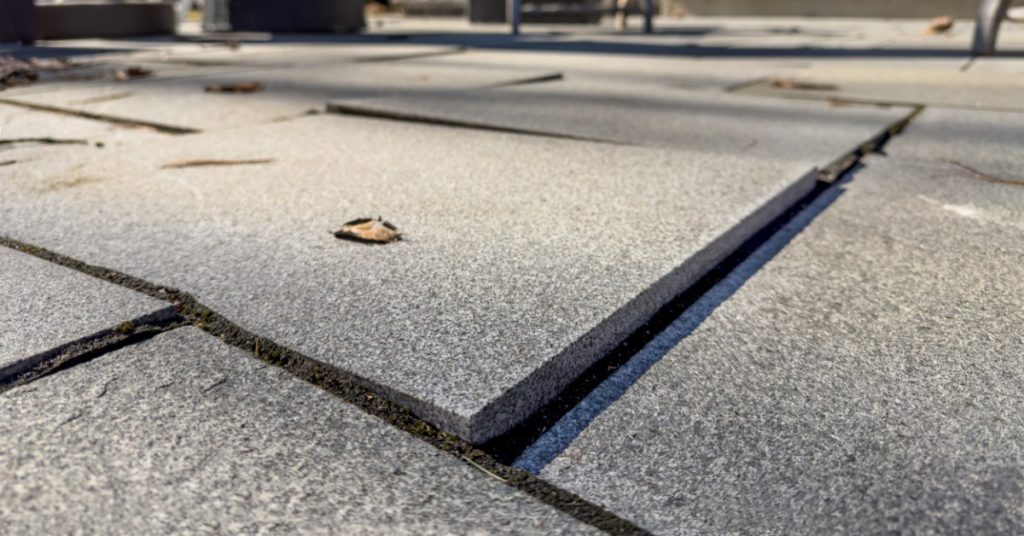 Sunlight shining on a residential patio made of large gray pavers. Several pavers are uneven and damaged.