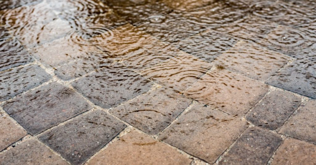 A large puddle pooling on patio pavers on a rainy day. Raindrops cause ripples in the puddle surface.