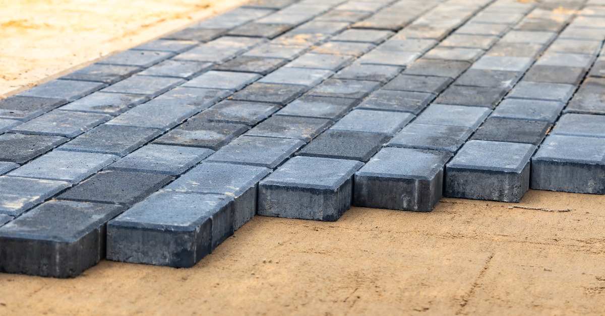 Sunlight beaming down on dark gray patio pavers partially installed outside. The pavers sit on a bed of sand.