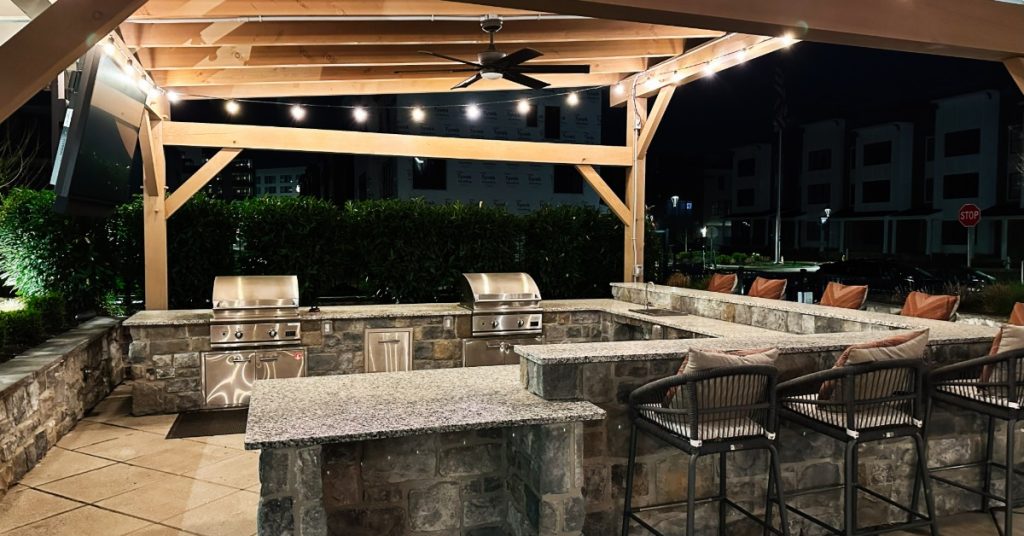 String lights hanging from a wooden arbor in a home’s backyard. A residential kitchen is built below the arbor.