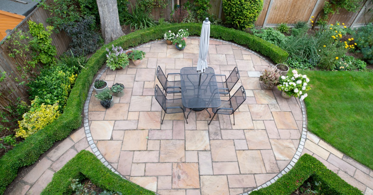 Bright sunlight shining on a home’s back patio. The patio has four chairs and a small table under an umbrella.