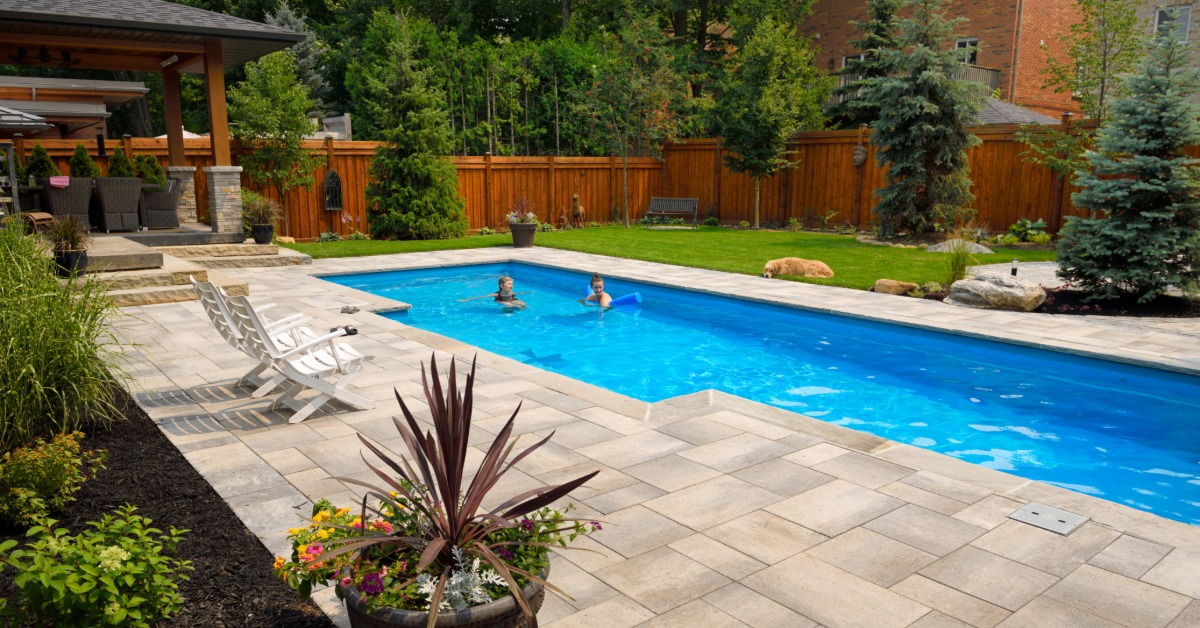 Two people in a long in-ground pool in a residential backyard during the daytime. Concrete pavers surround the pool.