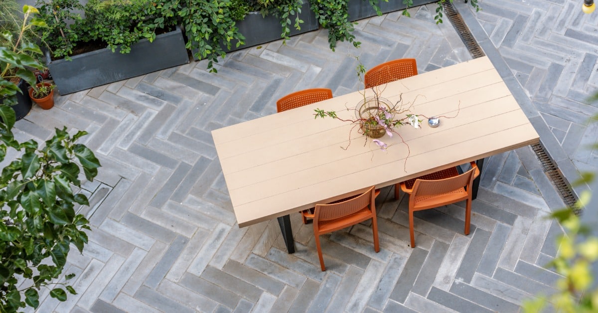 A long wooden table surrounded by four chairs on a patio made of concrete pavers arranged in a herringbone style.